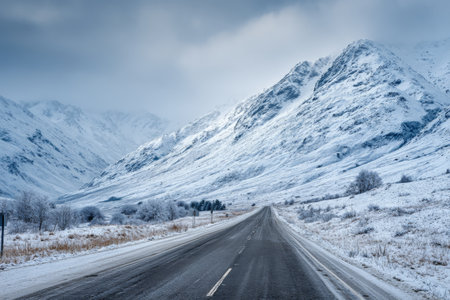 A winding road cuts through a breathtaking winter landscape, lined by majestic snow-covered mountains and a chilly, overcast sky. The serene scene captures the beauty of nature in winter.の素材