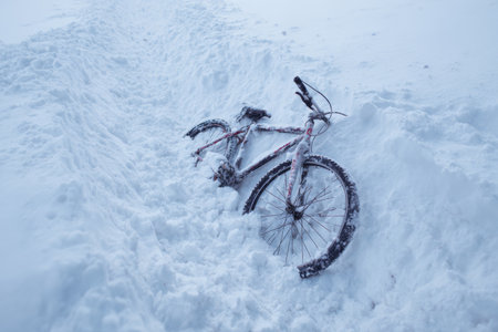 A bicycle lies partially buried in deep snow near a pathway. The scene shows a cold winter day with a clear view of a snowy landscape and subtle light.の素材