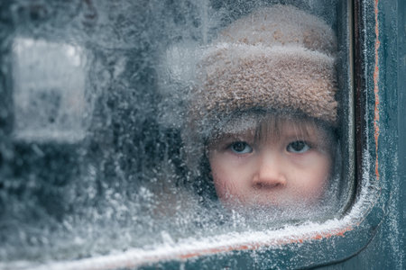 A young child gazes through a frosted window. The cold air creates a beautiful pattern of ice, while the child appears curious and contemplative in the chilly atmosphere.の素材