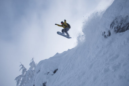 A snowboarder launches off a snowy cliff, surrounded by a winter landscape filled with fresh powder. The bright yellow jacket contrasts with the white snow under the cloudy sky.の素材