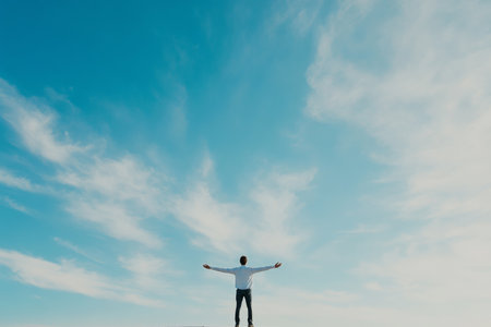 A person stands on a raised surface, arms stretched wide, embracing the tranquility of a clear blue sky with soft, wispy clouds on a peaceful afternoon.の素材