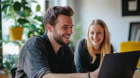 A young man and woman share a joyful moment while working collaboratively on a laptop in a bright workspace surrounded by greenery, showing their strong friendship and teamwork.の素材