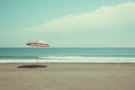 A striped beach umbrella stands alone on the sandy beach, providing shade against the sun while gentle waves lap at the shore. The ocean glistens under the bright sky.の素材