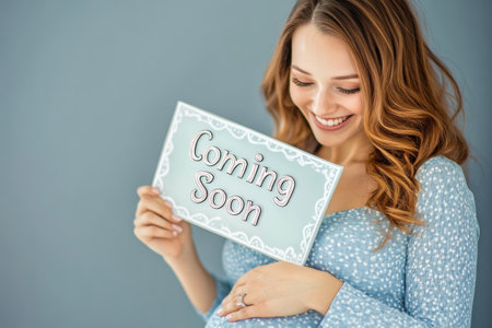 A happy pregnant woman poses with a sign reading Coming Soon, cradling her baby bump. Her cheerful expression and a gentle background create a warm atmosphere celebrating new life.の素材