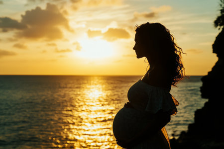 A serene silhouette of a pregnant woman stands by the ocean during sunset. The sky is painted with soft golden hues, creating a peaceful and reflective atmosphere.の素材