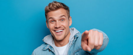 A cheerful man smiles broadly while pointing at the camera. He wears a denim jacket over a white shirt, set against a vibrant blue background. His joyful expression captures attention.の素材