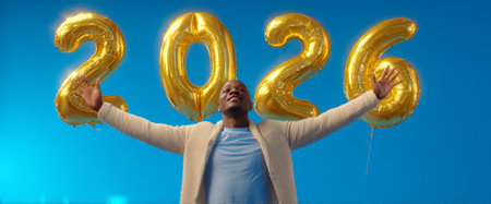 A person joyfully celebrates, holding giant gold balloons that display the year 2026. The bright blue background enhances the festive atmosphere of the moment.の素材