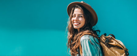 A woman with long hair and a brown hat is smiling cheerfully. She stands against a vibrant blue wall, wearing a casual outfit and carrying a backpack. Sunlight highlights her joy.の素材