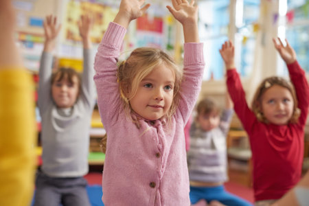 A group of children is engaging in a yoga session in a colorful classroom. They are stretching with their hands raised, enjoying the activity and focused on their instructor.の素材
