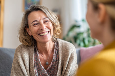 Two women are sitting together on a couch in a bright, cozy living room. They are engaged in a warm conversation, sharing smiles and laughter, creating a relaxed atmosphere.の素材