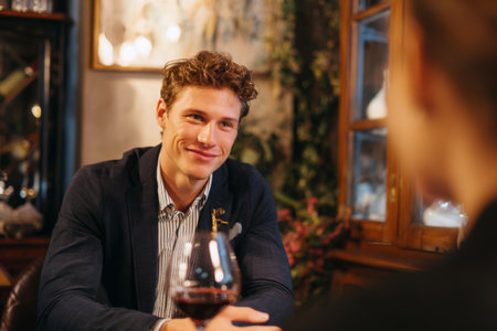 A young man smiles while engaging in conversation with another person across the table in a warm, inviting restaurant. Wine glasses sit on the table, enhancing the relaxed atmosphere.の素材