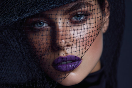 A model showcases bold violet lipstick and captivating eye makeup while wearing a black netted headpiece. The dramatic contrast draws attention to her striking features.の素材