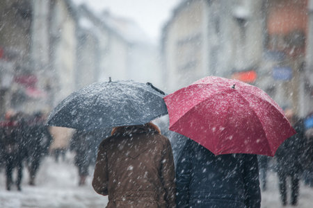 A couple sheltered by umbrellas walks closely together on a snowy city street. Snowflakes fall heavily around them, and people bustle in the background, creating a lively winter atmosphere.の素材