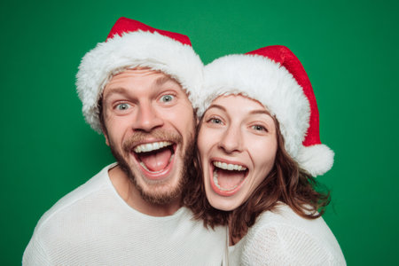 A couple expresses joy and happiness while wearing fluffy red and white Santa hats. They stand closely together against a vibrant green backdrop, celebrating the festive season.の素材