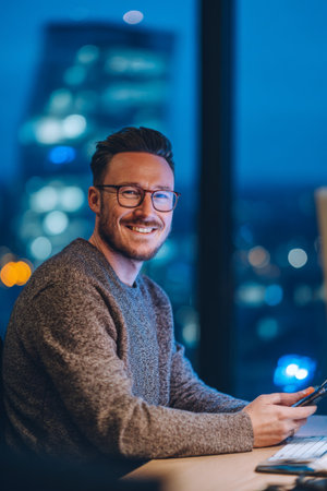 A man with glasses sits at his desk, smiling while holding his phone. The background shows glowing city lights as evening sets in through the window.の素材