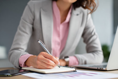 A woman in a business suit writes notes in a notepad while sitting at a desk. Her laptop is open and documents are spread out in a contemporary office environment.の素材