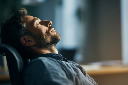 A man with a beard leans back in an office chair with his eyes closed. Sunlight filters through the window, creating a calm atmosphere. He seems to be enjoying a moment of peace in his workspace.の素材