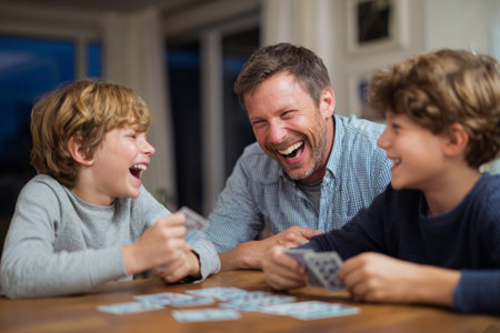 A man and two boys are gathered around a table, joyfully playing cards. Laughter fills the room as they share a warm evening together, enjoying their time.の素材