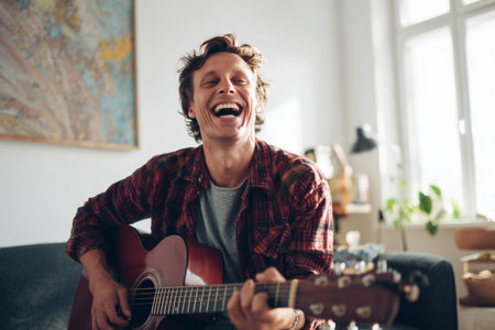 A happy man is playing his acoustic guitar in a bright, inviting room. He smiles widely, enjoying the music and creating a cheerful atmosphere. Green plants and art add warmth to the space.の素材