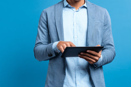 A professional man wearing a light blue shirt and gray blazer focuses on a tablet in his hands. He stands in front of a solid blue wall, showing a modern workspace feel.の素材