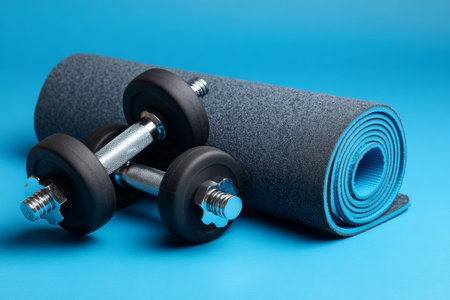 Two dumbbells sit beside a rolled exercise mat on a blue surface. The scene is set for a workout session, showcasing essential fitness gear and inviting activity.の素材