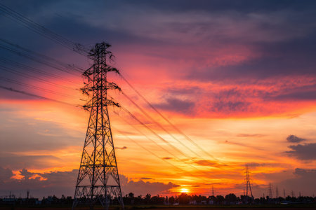 A sunset displays bright colors in the sky behind tall power lines. The landscape shows a mix of clouds and an open field. The scene is near a city during evening hours.の素材
