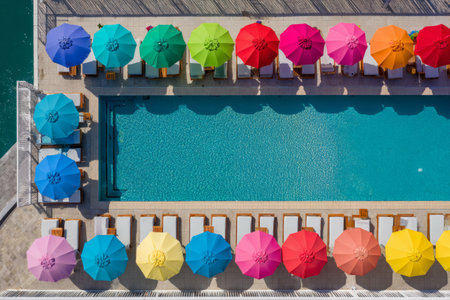 Colorful umbrellas surround a pool at a beach club hotel. This aerial view shows the layout of sun loungers beside the clear water on a sunny day.の素材