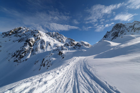 A winding trail cuts through deep snow on a mountain landscape. Majestic peaks rise around, with a bright blue sky overhead showcasing a beautiful winter day.の素材