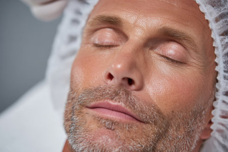 A 50 year old man relaxes with closed eyes while receiving an anti-aging facial treatment at a modern clinic. He enjoys high-quality skin care in this close-up scene.の素材