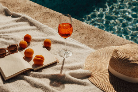Bright light shines on a white towel by the pool. Whole and half-eaten apricots are present, along with a drink, sunglasses, a hat, and an open notebook with notes.の素材