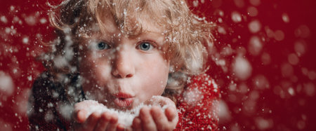 A cheerful child is blowing snowflakes from their hands, surrounded by a flurry of snow against a bright red background. The joyful moment captures the spirit of winter and play.の素材