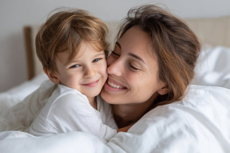 A mother and her young son smile as they embrace in bed, wrapped in soft blankets. The atmosphere is warm and loving, creating a sense of comfort and happiness.の素材