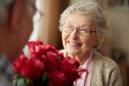 A joyful elderly woman beams as she receives a beautiful bouquet of red flowers from a loved one in a warm and invitingの素材