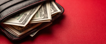 A wallet made of black leather reveals several neatly arranged dollar bills. The wallet rests on a solid red background, creating a striking visual contrast.の素材