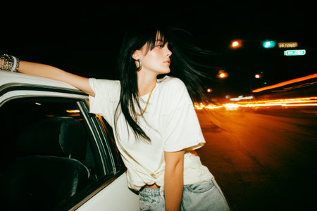 A 25-year-old woman leans out of a white car window with long hair flowing in the night wind. Her jewelry shines as the car speeds down the road, creating a sense of motion.の素材