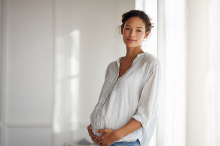A pregnant woman stands in a bright space. She gently touches her belly while wearing casual clothes. The warm light creates a soft atmosphere around her.の素材