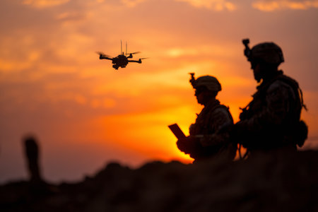 Silhouettes show two soldiers using a laptop and observing a drone flying in the sky. The scene is set during a scouting operation at sunset.の素材