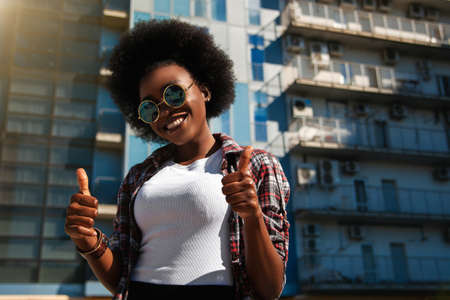Image of a happy optimistic emotional young american african student posing over city background dressed casual and wearing sunglasses, showing thumbs up gesture. Lifestyle.の写真素材