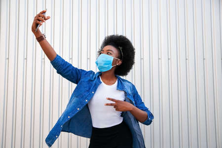 Health, pandemic and safety concept - young black woman with black power hair wearing protection mask making a selfie on grey background in city.の写真素材