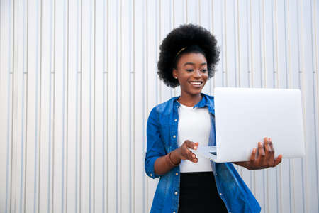 Close-up portrait cute smiling African American young woman with dark curly hair holding in hands laptop and looking at him, over gray background in city, empty copy space. Distant online education with tutor.の写真素材