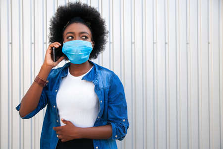 Medicine healthcare concept - young american african woman, dressed casual, wearing disposable medical protective mask using smart phone in the city on gray background. Copy space.の写真素材
