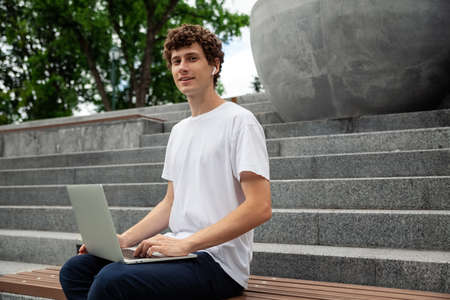 European young male in wireless earphones wearing white t-shirt sitting in a city park and looking in smartphone. Freelance work concept outdoors. Empty copy space.elance work concept outdoors.の写真素材