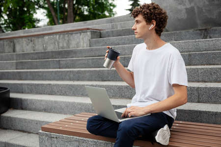 European young male in wireless earphones wearing white t-shirt sitting in a city park and looking in smartphone. Freelance work concept outdoors. Empty copy space.elance work concept outdoors.の写真素材