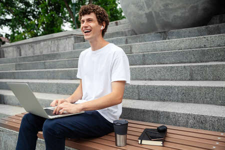 European young male in wireless earphones wearing white t-shirt sitting in a city park and looking in smartphone. Freelance work concept outdoors. Empty copy space.elance work concept outdoors.の写真素材