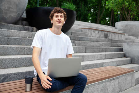 European young male in wireless earphones wearing white t-shirt sitting in a city park and looking in smartphone. Freelance work concept outdoors. Empty copy space.elance work concept outdoors.の写真素材