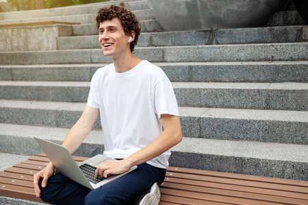 European young male in wireless earphones wearing white t-shirt sitting in a city park and looking in smartphone. Freelance work concept outdoors. Empty copy space.elance work concept outdoors.の写真素材