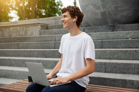 European young male in wireless earphones wearing white t-shirt sitting in a city park and looking in smartphone. Freelance work concept outdoors. Empty copy space.elance work concept outdoors.の写真素材
