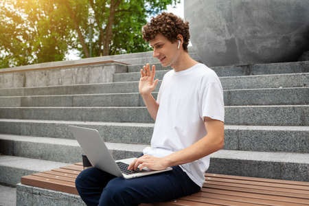 European young male in wireless earphones wearing white t-shirt sitting in a city park and looking in smartphone. Freelance work concept outdoors. Empty copy space.elance work concept outdoors.の写真素材