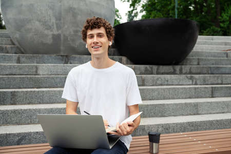 European young male in wireless earphones wearing white t-shirt sitting in a city park and looking in smartphone. Freelance work concept outdoors. Empty copy space.elance work concept outdoors.の写真素材