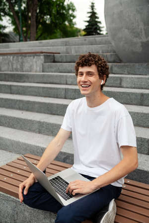 European young male in wireless earphones wearing white t-shirt sitting in a city park and looking in smartphone. Freelance work concept outdoors. Empty copy space.elance work concept outdoors.の写真素材